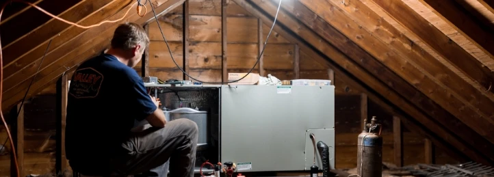 Technician servicing an HVAC unit inside an attic with exposed wooden beams and ductwork.